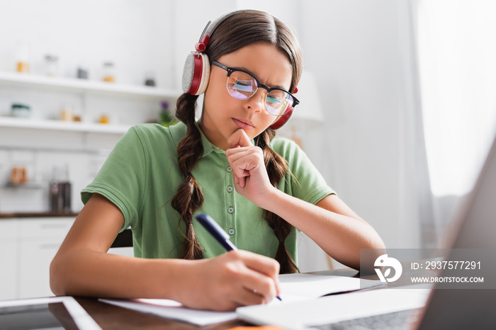 Thoughtful hispanic girl writing in copy book, while sitting at table with laptop in kitchen on blur