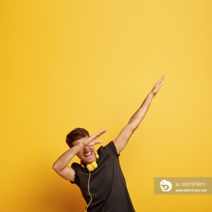 Cheerful young man makes dab dance gesture, shows dabbing movement, has fun in club, uses modern hea