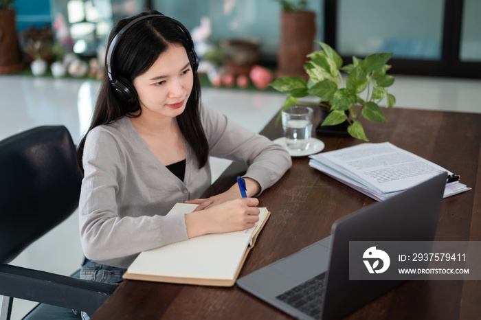 Young asian girl student wears wireless headphones write on the notebook to study language online wa