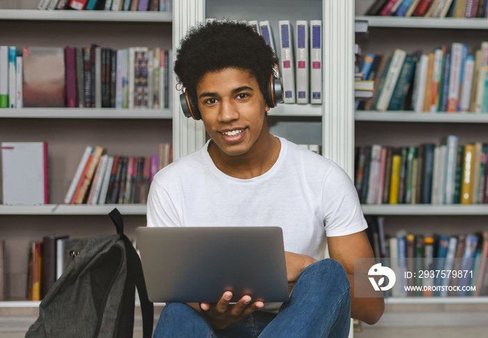 非裔美国学生，头戴耳机，靠在书架上，使用笔记本电脑