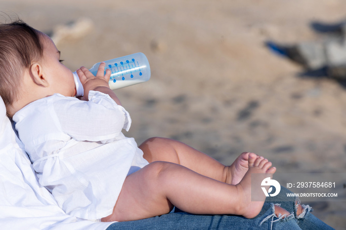 Newborn drinking milk from a baby bottle, sitting on mom legs outdoors