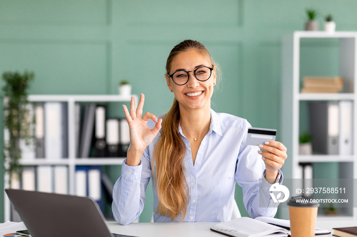 Accountant banker lady holding credit card and showing ok sign while sitting at workplace, smiling a
