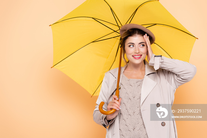 joyful woman in fashionable autumn outfit touching face under yellow umbrella on peach