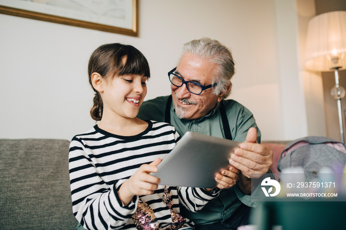 Smiling grandfather and granddaughter looking at digital tablet while sitting in living room at home