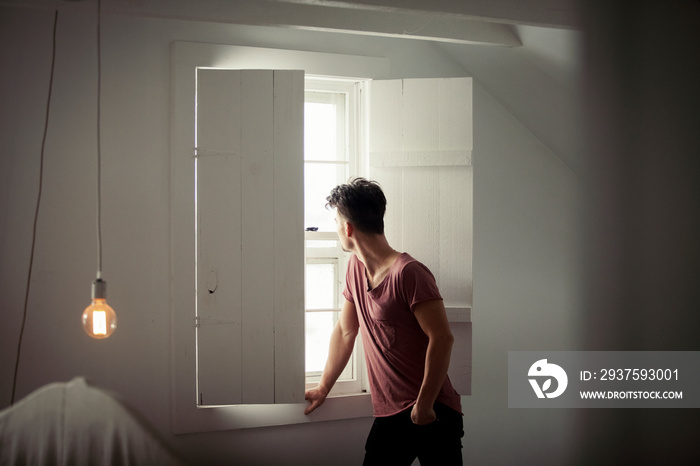 Thoughtful man with hand in pocket looking through window while standing at home