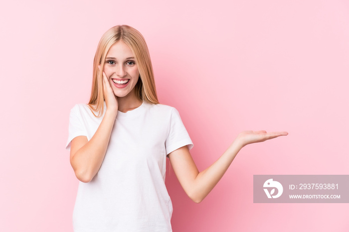 Young blonde woman on pink background holds copy space on a palm, keep hand over cheek. Amazed and d