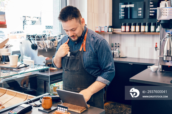 Young bearded Caucasian male man cashier barista in restaurant cafe at work. Seller using touch pad 