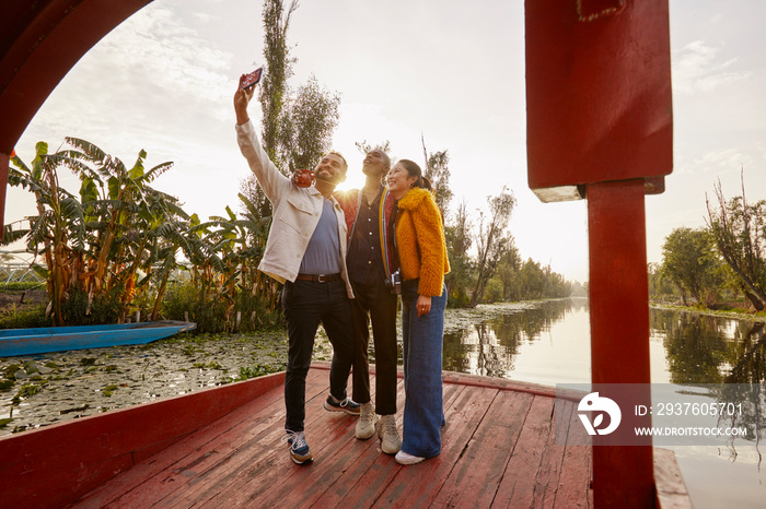 Smiling young friends taking selfie with smartphone while standing on trajinera boat