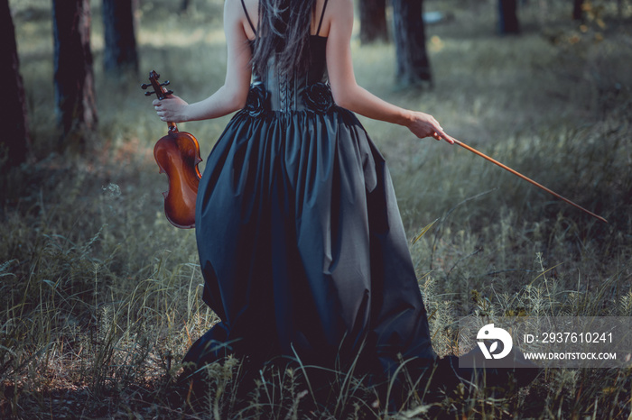 back view of woman in witch costume walking on forest, holding violin