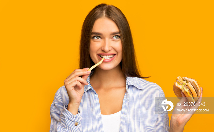 Happy Girl Eating Burger And French Fries Standing, Yellow Background