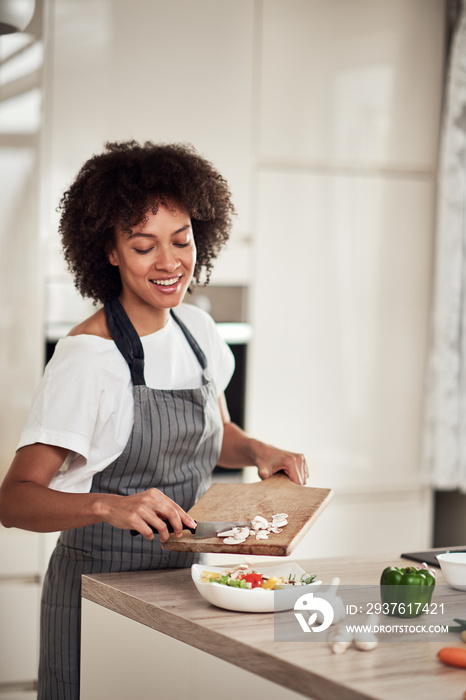 微笑的混血女人穿着围裙，把切碎的蘑菇和蔬菜一起放在碗里。厨房内部