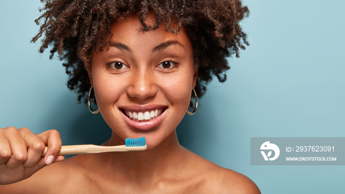 Beautiful dark skinned lady with snow white smile, brushes teeth, holds toothbrush, has Afro haircut