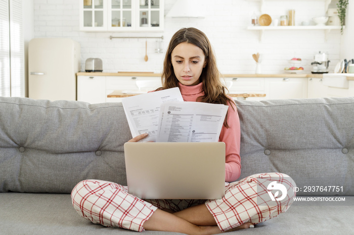 Young girl siiting on sofa in living room with laptop open and bills checking details of payment