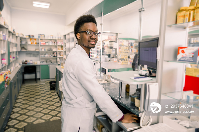 Portrait of young smiling African male pharmacist working with computer behind counter in pharmacy