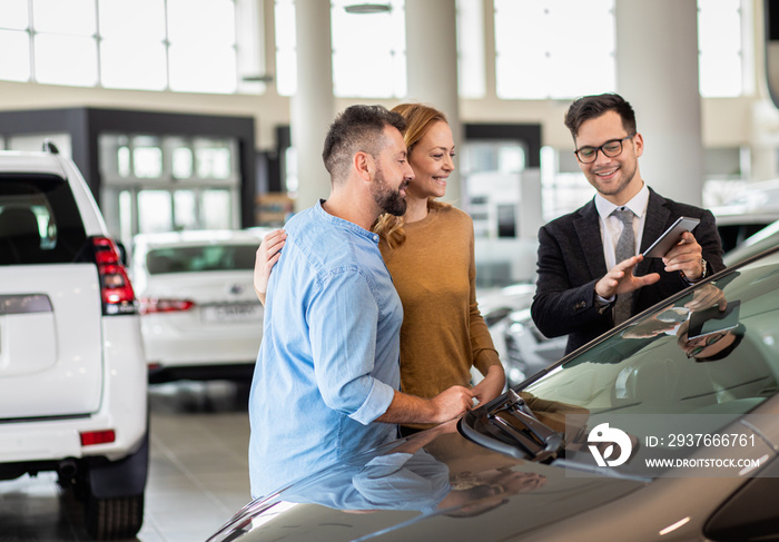 Young car salesman showing to middle age couple new automobile at dealership salon.