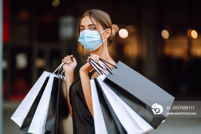 Stylish woman in black dress in protective medical mask holding shopping bags near center mall. Shop