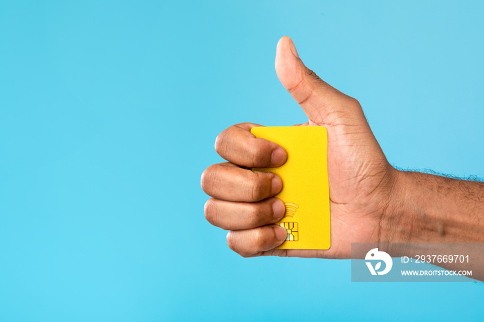 千禧一代黑人拿着信用卡，在蓝色工作室背景上竖起大拇指的裁剪图