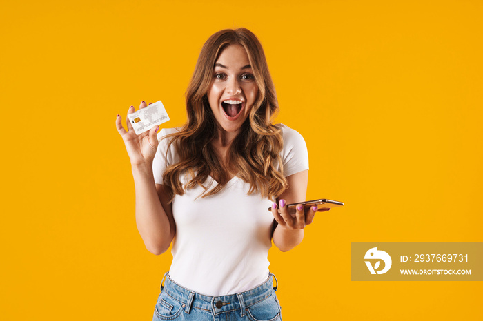 Photo of pretty young woman rejoicing while holding credit card and smartphone isolated over yellow 