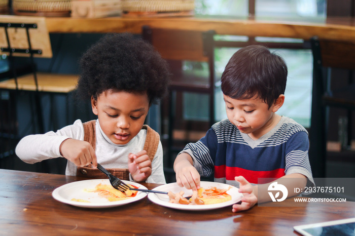 Cute African American boy with curly and adorable Asian kid eating meal at the table indoor, happy c