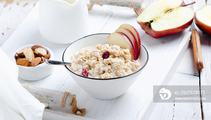 Oatmeal porrige with red apple in ceramic bowl over rustic white wooden tray.