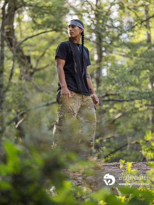 一个留着长辫子的年轻土著男子站在森林里的原木上的垂直图像