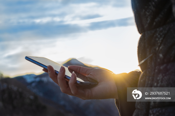 日落时，在阳光、山脉和多云的蓝光下，一位女士拿着智能手机在户外的特写