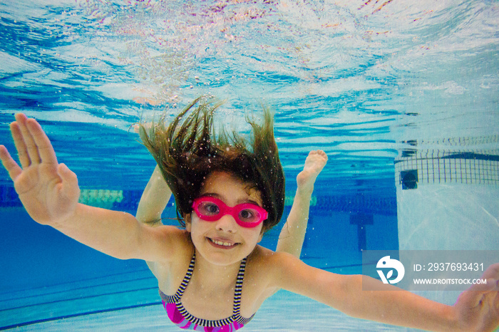 Girl underwater in swimming pool wearing goggles looking at camera smiling