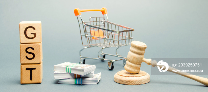 Wooden blocks with the word GST, money and a supermarket trolley with a judge’s gavel. Tax, which is imposed on the sale of goods and services